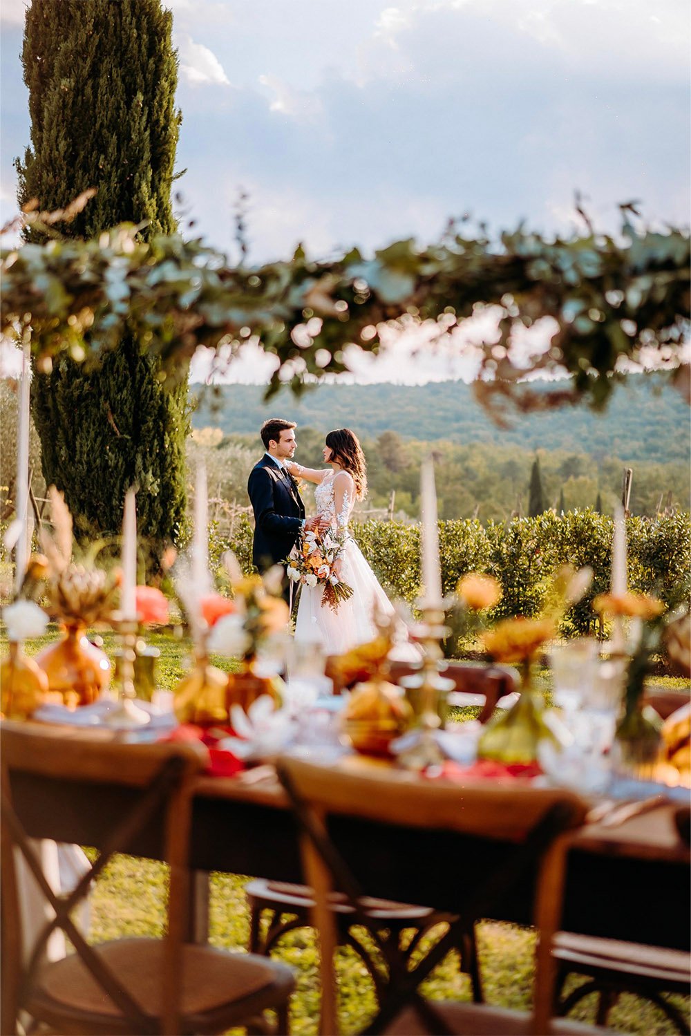 Borgo Iesolana, Tuscany, Italy — a newlywed couple standing together among cypress trees and rolling Tuscan hills, photographed through softly blurred floral arrangements in an editorial wedding photography style.