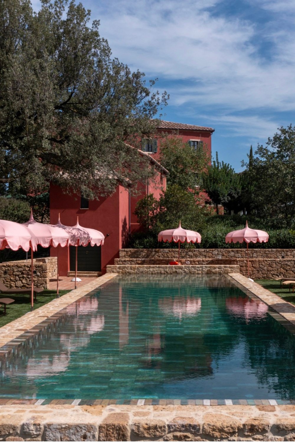 Casa Newton, Tuscany, Italy — a striking outdoor swimming pool surrounded by stone walls, a terracotta villa, and pink parasols, photographed in a vibrant editorial style.
