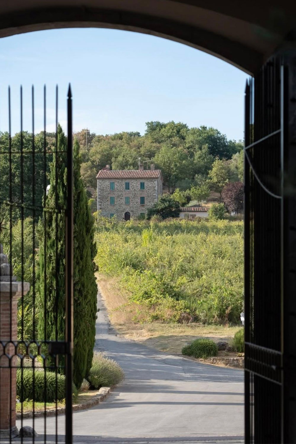 Castel Monastero, Tuscany, Italy — a view through a historic stone archway and wrought-iron gate toward the surrounding countryside and cypress-lined driveway, captured in an editorial style.