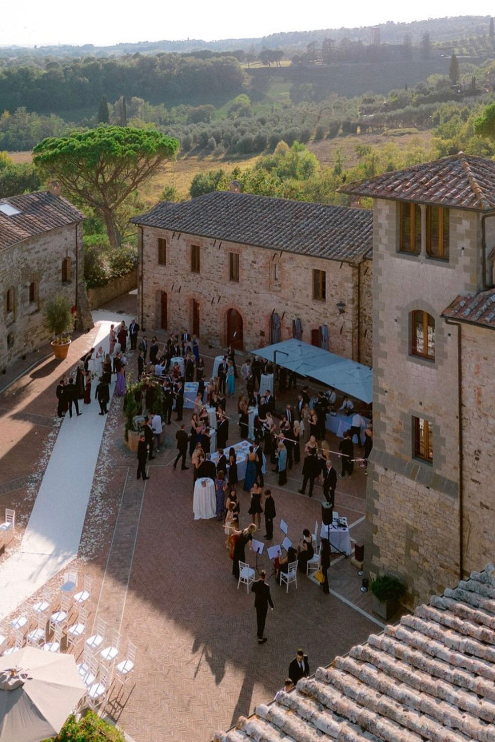 Castel Monastero, Tuscany, Italy — an elevated view of a wedding reception taking place in the historic stone courtyard, with elegant tables, guests, and a white ceremony aisle captured in an editorial style.