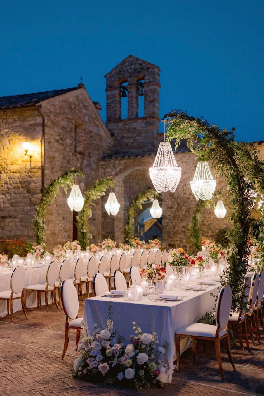 Castel Monastero, Tuscany, Italy — An elegant outdoor wedding reception at Castel Monastero in Tuscany, with candlelit tables, floral arrangements and crystal chandeliers set against the historic stone architecture in a romantic editorial style.