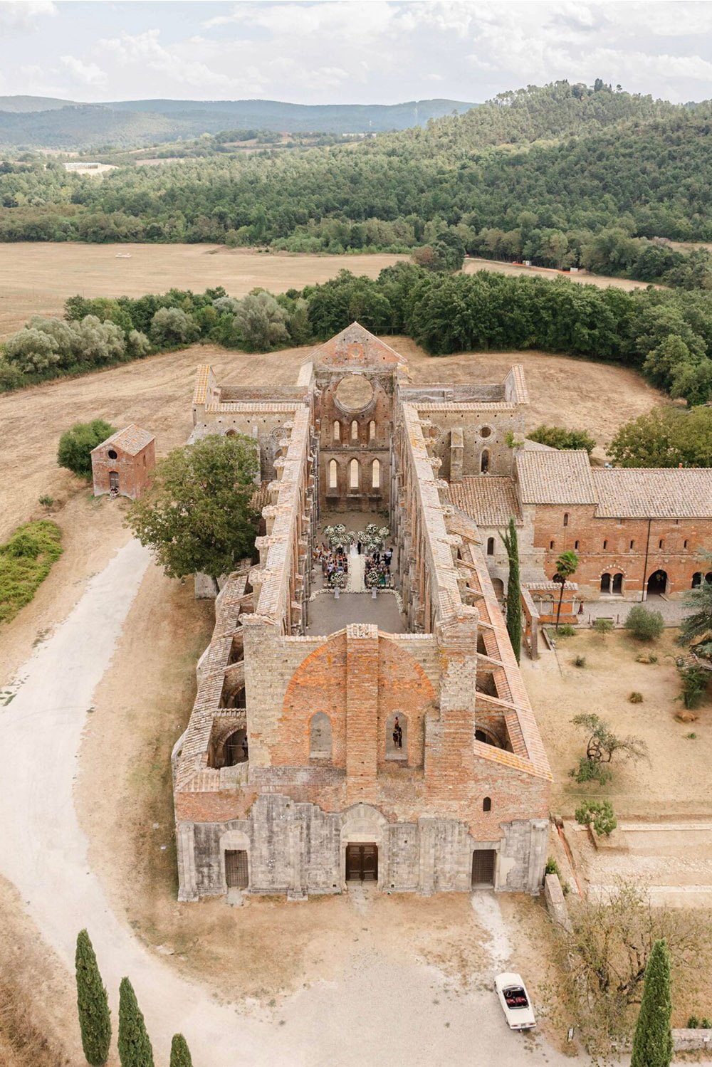 San Galgano Abbey wedding venue: Aerial view of a wedding ceremony inside the historic San Galgano Abbey in Tuscany, Italy, surrounded by rolling countryside in a romantic editorial style.